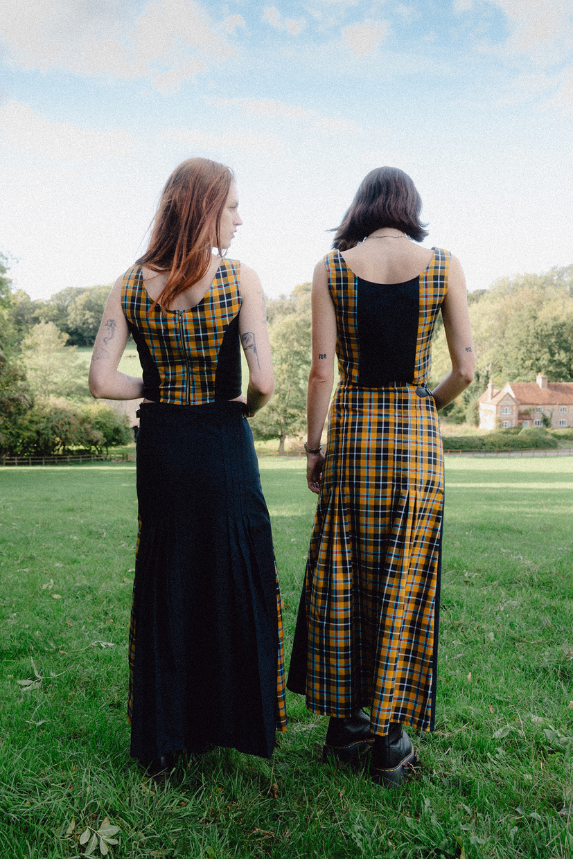 Two women wearing matching black and yellow checkered dresses standing in a grassy field.