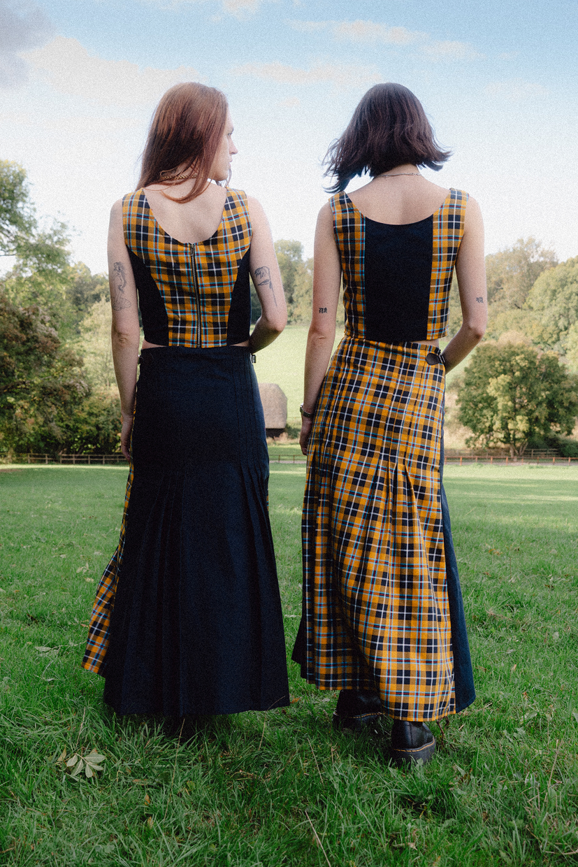 Two women wearing matching black and yellow checkered dresses standing in a grassy field.